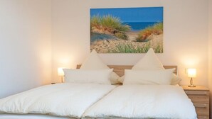 A double bed with white bedding, two bedside lamps, and a painting of a beach scene on the wall. - Residenz zum Südstrand - Kutte (Borkum)