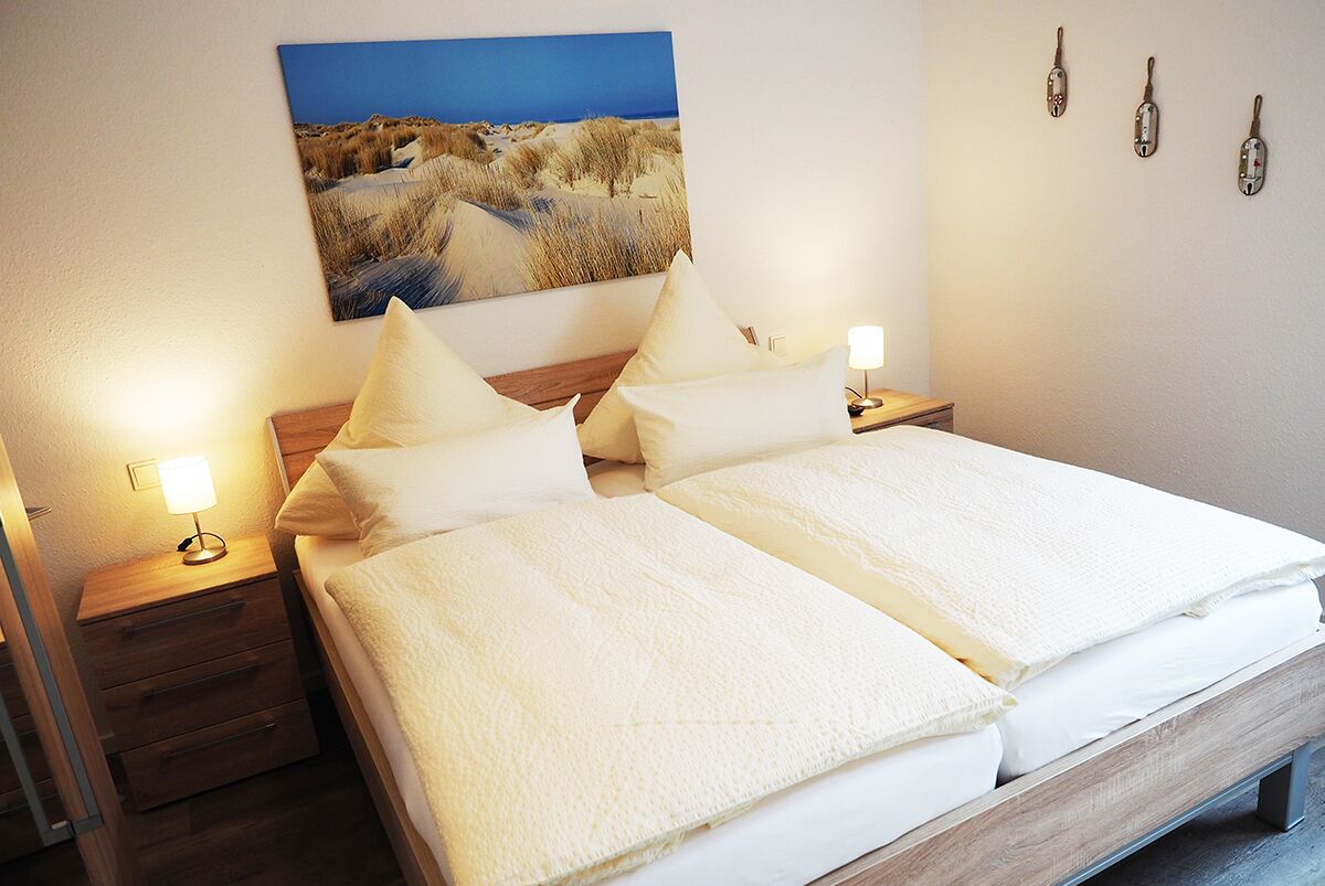 Een tweepersoonsbed met witte beddengoed, twee nachtkastjes met lampen, een aan de muur hangend schilderij van een strand en drie haken aan de muur.
