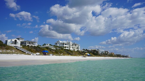 A beachfront property with multiple white buildings, a sandy beach, and a clear blue sky with clouds. - Seaside, FL Beach Home - Close to Pool & Beach; + 2 Adult Bikes! (Santa Rosa Beach)