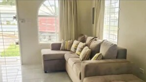 A living room with a beige sectional sofa, a small ottoman, and a large window with curtains. - Remarkable 2-bed House in Discovery Bay (Discovery Bay)