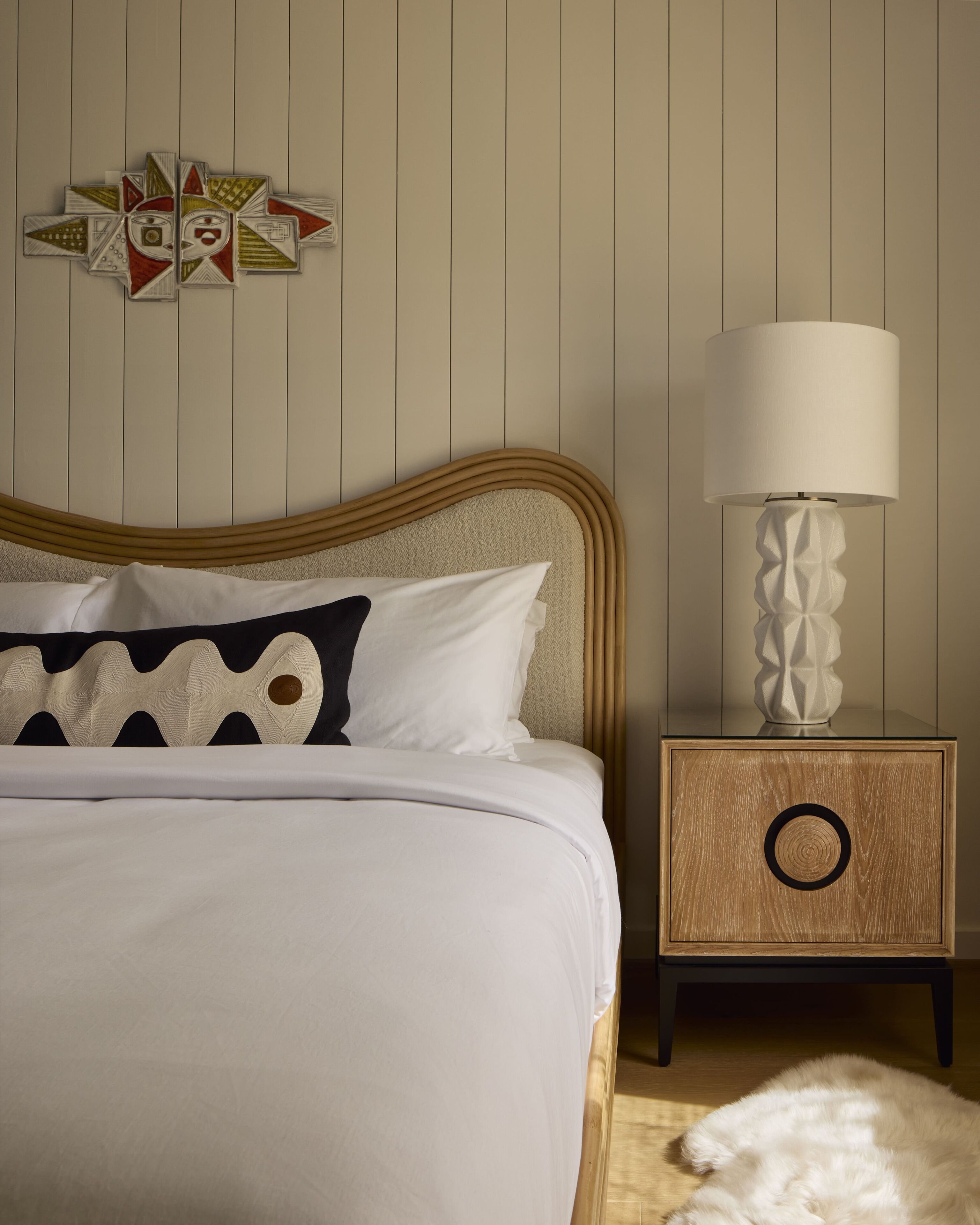 A neatly made bed with white linens and a patterned pillow, a wooden bedside table with a lamp, and a wall with a geometric wall art piece.