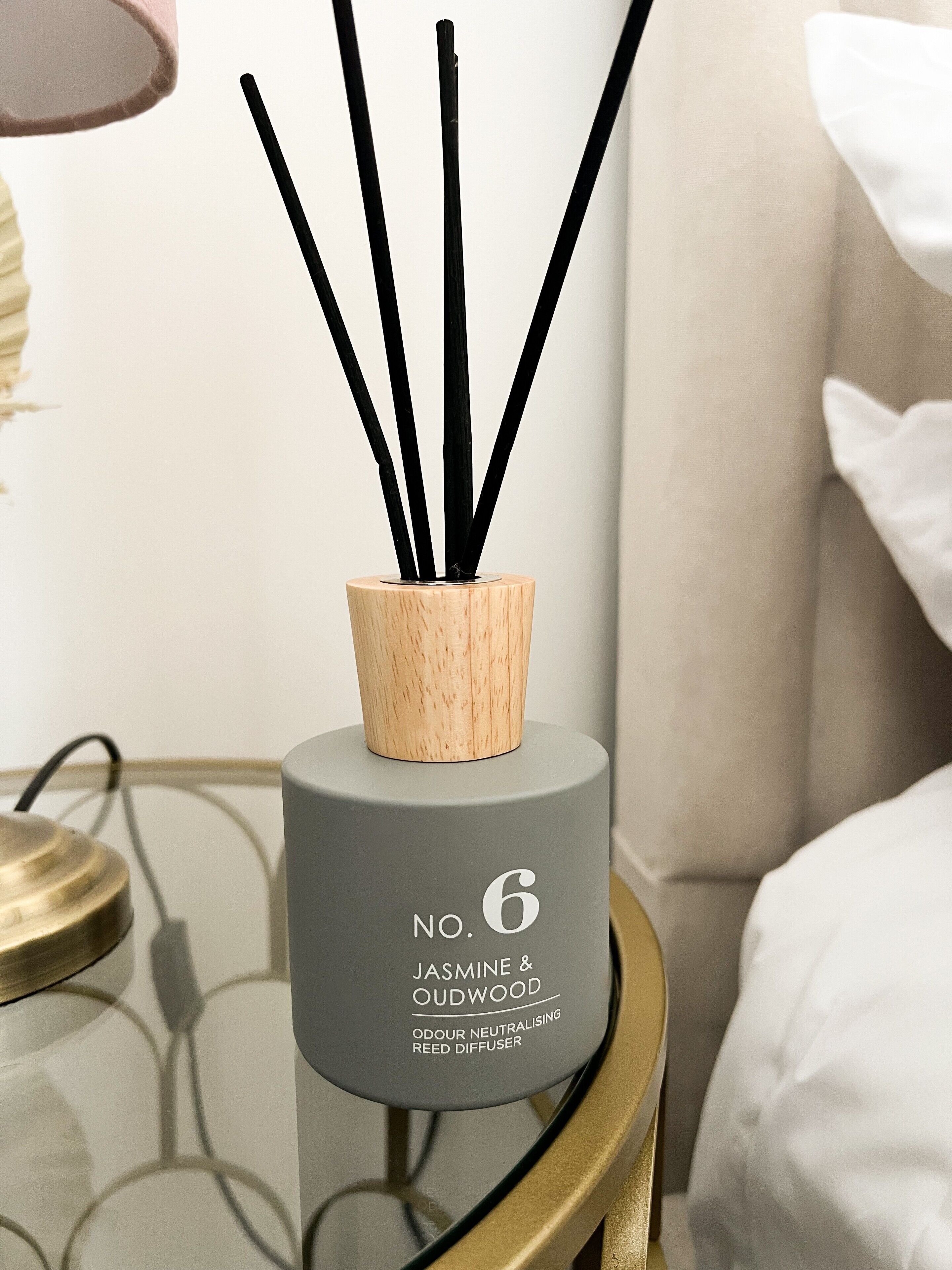 A grey diffuser with black reeds labeled 'Jasmine & Oudwood' on a bedside table.