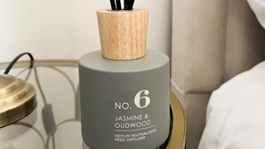 Harmaa diffuusori, jossa on mustia tuoksutikkuja, ja sen etikettiin on painettu teksti ”Jasmine & Oudwood”. Se on sijoitettu yöpöydälle.