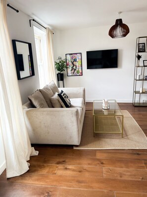 Ein Wohnzimmer mit einer Couch, einem Glastisch, einem Fernseher, einer Pflanze und einem Bücherregal.