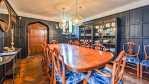 A dining room with a large wooden table and chairs, a chandelier, and a bookshelf filled with dishes.