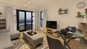 Un salon moderne avec un canapé, un pouf, une table à manger en verre et un téléviseur.