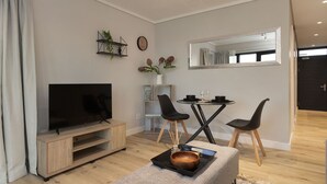 Un salon moderne doté d’un téléviseur à écran plat, d’un meuble de rangement en bois et d’un coin repas avec deux chaises noires et une petite table.