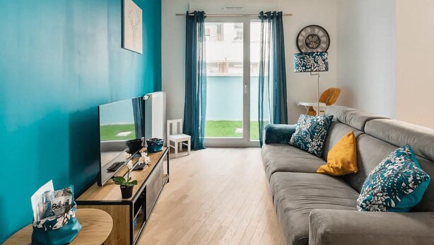 Una sala de estar moderna con un sofá gris, paredes verde azulado, un televisor y un balcón con cortinas azules.