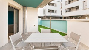 Un patio con una mesa y sillas blancas, un toldo color turquesa y vistas de un edificio residencial con balcones.