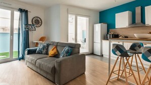 Una sala moderna con un sofá grisáceo, cojines estampados y piso de madera. Se ve una zona de cocina con armarios blancos y una pared azulada.