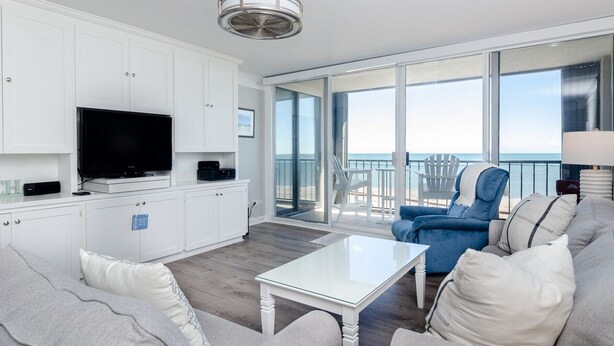 A modern living room with a flat-screen TV, white cabinets, a glass coffee table, and a balcony with a view of the ocean. - Fantastic upgraded Station One condo (Wrightsville Beach)