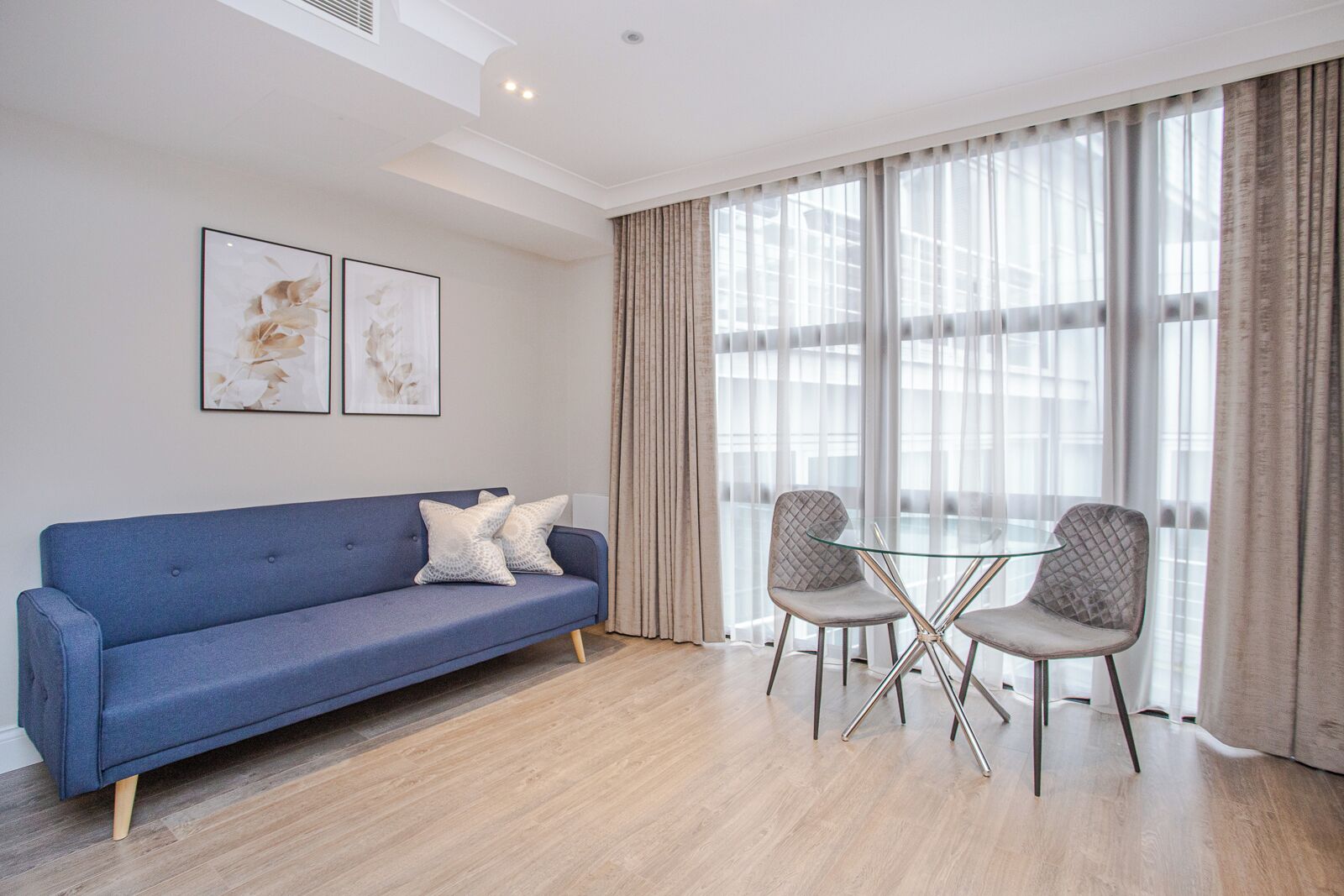 Een moderne woonkamer met een blauwe bank, twee grijze stoelen, een glazen tafel en grote ramen met transparante gordijnen.