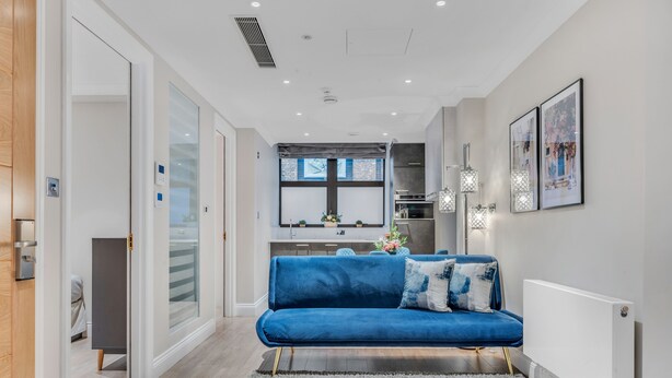 A modern living room with a blue sofa, a grey rug, and a kitchen in the background. - Cleveland Residences Farringdon (London)