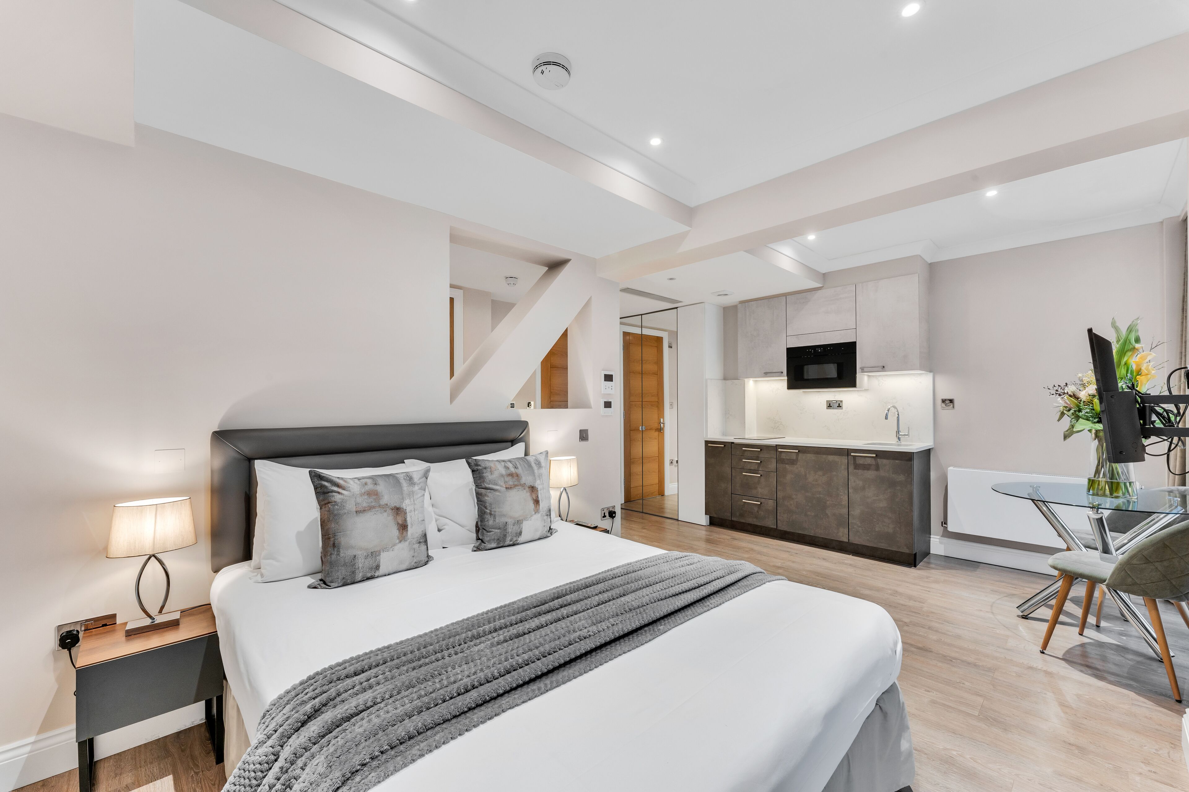 A modern hotel room with a large bed, bedside tables, a small desk, and a kitchenette.