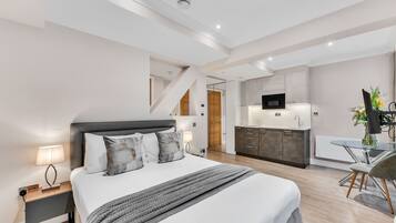 A modern hotel room with a large bed, bedside tables, a small desk, and a kitchenette.
