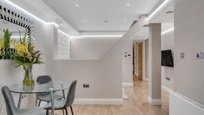 A modern hotel room with a glass dining table, grey upholstered chairs, a vase with flowers, a large mirror, and a flat-screen TV mounted on the wall. - Cleveland Residences Farringdon (London)