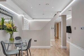A modern hotel room with a glass dining table, grey upholstered chairs, a vase with flowers, a large mirror, and a flat-screen TV mounted on the wall. - Cleveland Residences Farringdon (London)