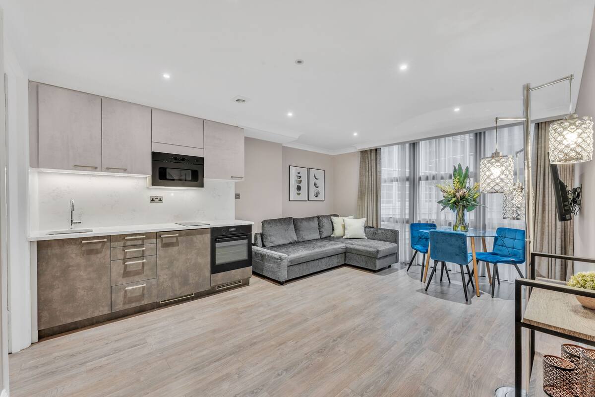 A modern kitchen with built-in appliances, a grey sectional sofa, blue chairs, and a dining table with a vase of flowers.