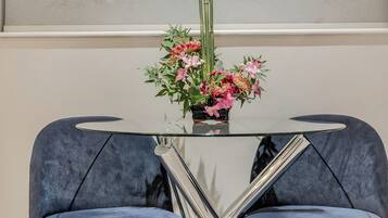 Ein moderner Esstisch mit Glasplatte und Metallbeinen, umrahmt von zwei blauen, gepolsterten Stühlen mit Holzbeinen. Auf dem Tisch steht eine Vase mit rosa Blumen.
