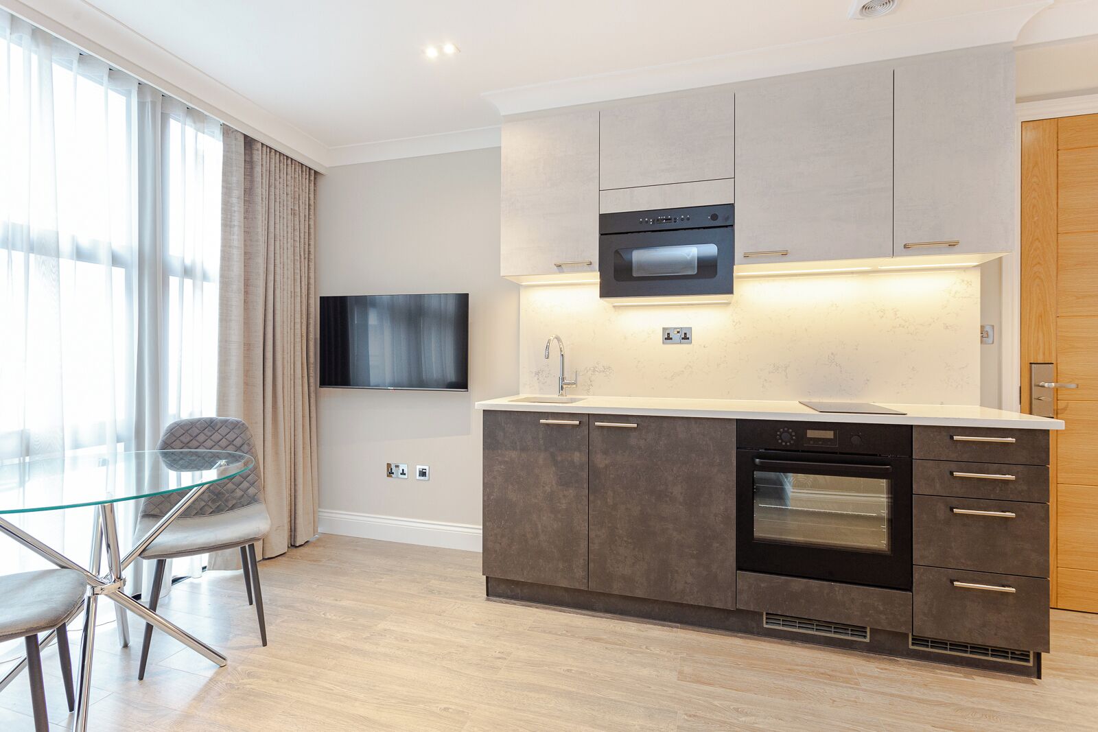 A modern kitchen with a flat-screen TV, built-in oven, and a dining area with a glass table and chairs.