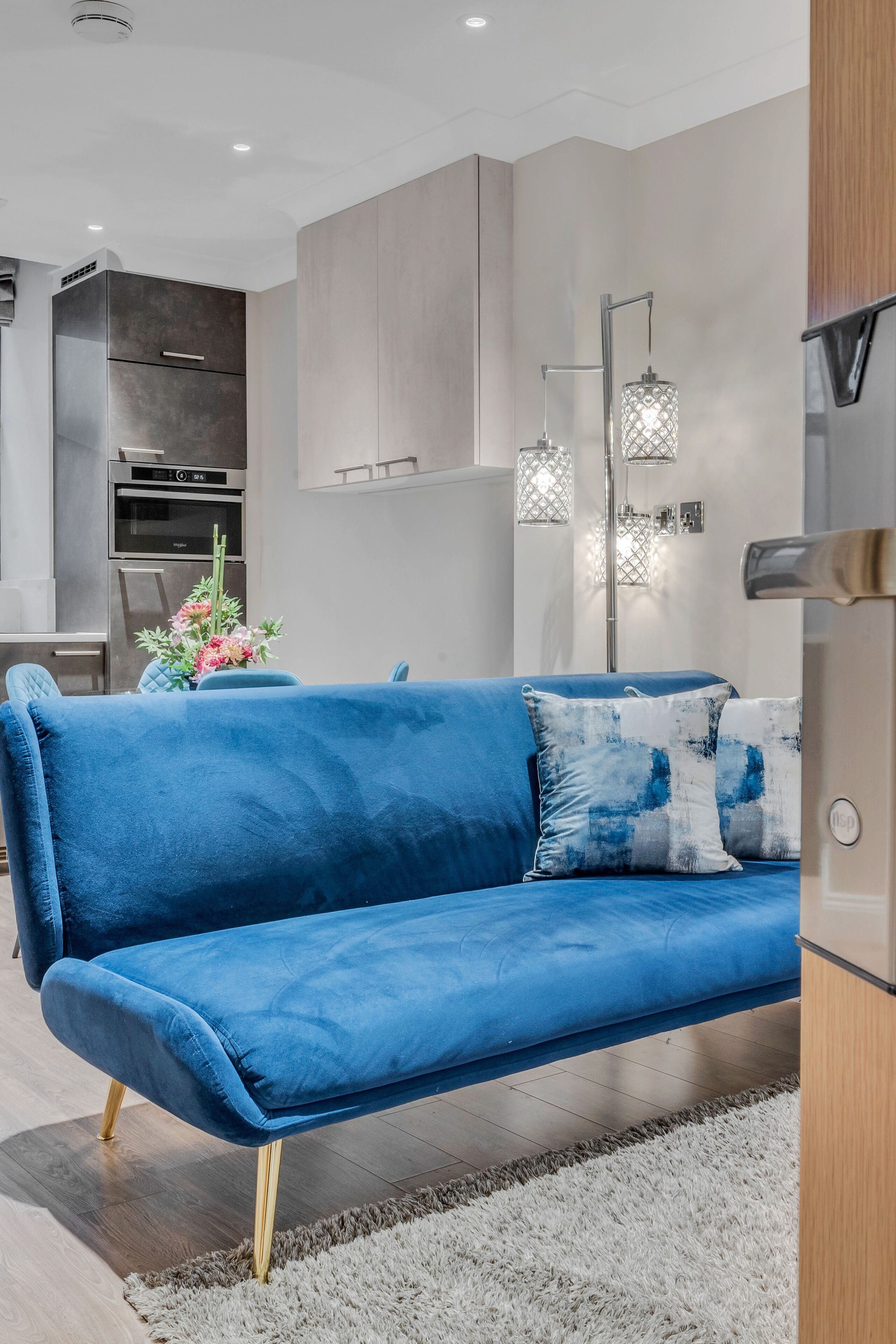 A modern living room with a blue sofa, a white rug, and a kitchen in the background.