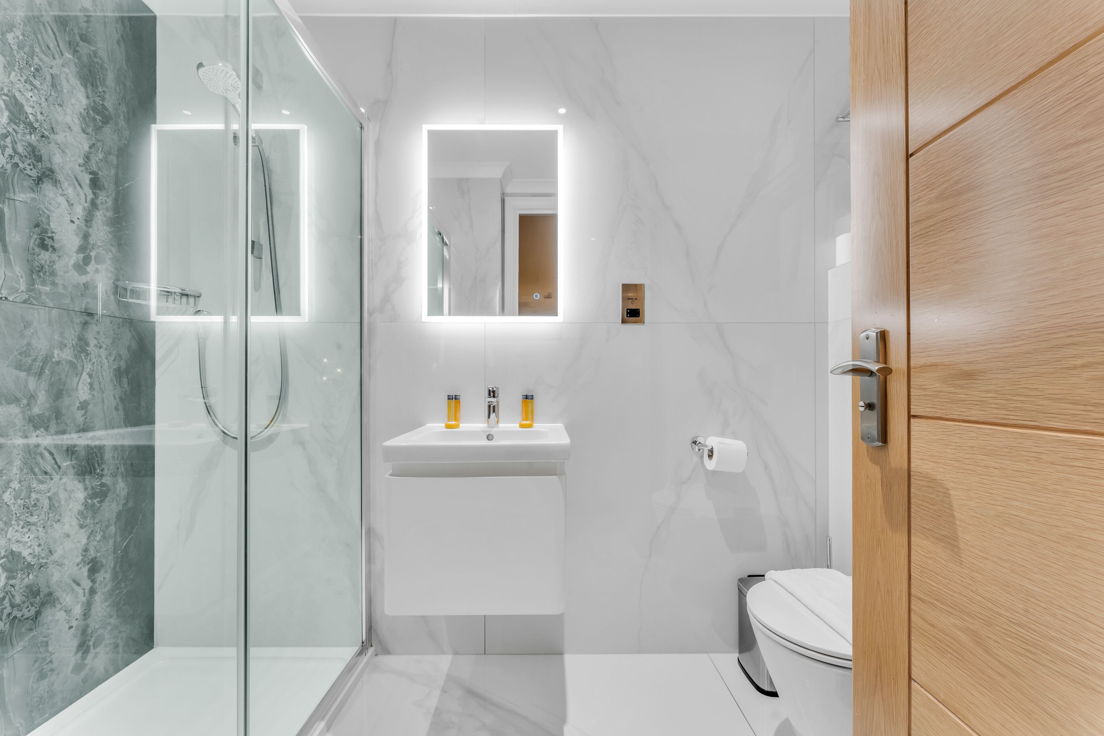 A modern bathroom with a glass-enclosed shower, a white sink with two bottles, a mirror with integrated lighting, and a wooden door.