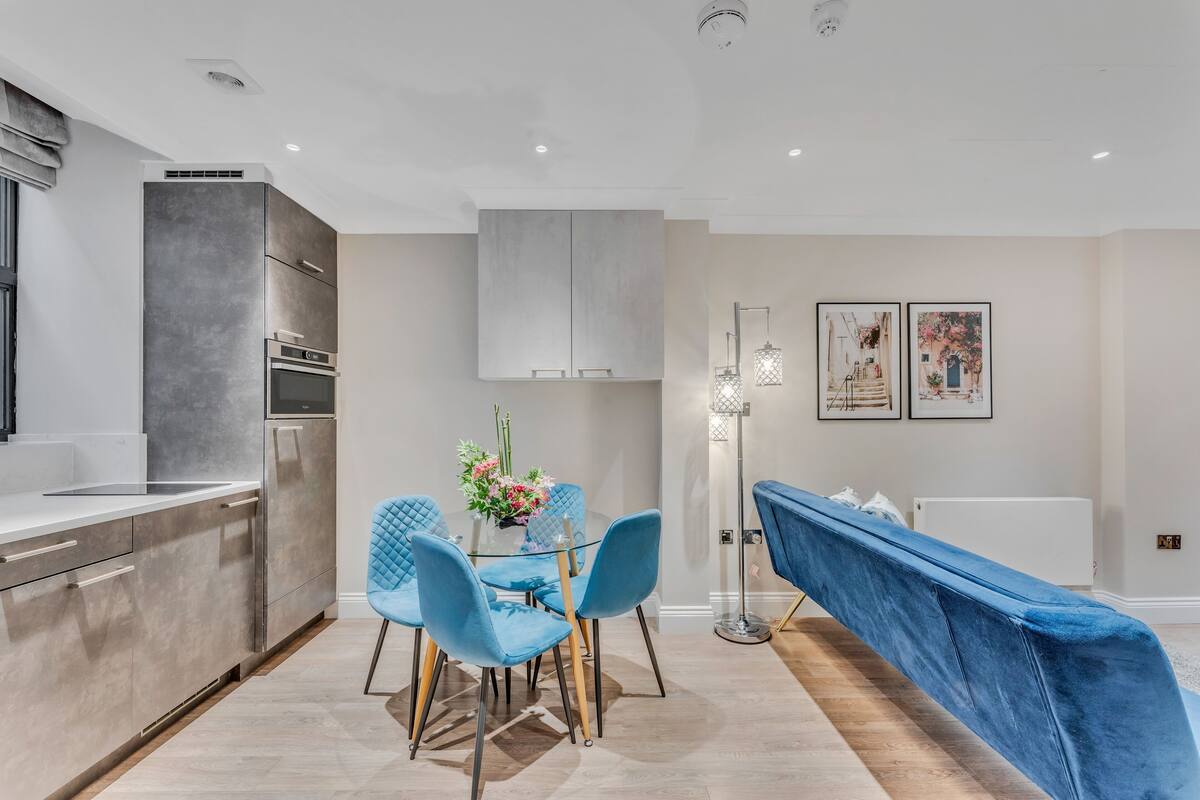 A modern kitchen with grey cabinets, a built-in oven, and a dining area with a glass table and blue chairs.
