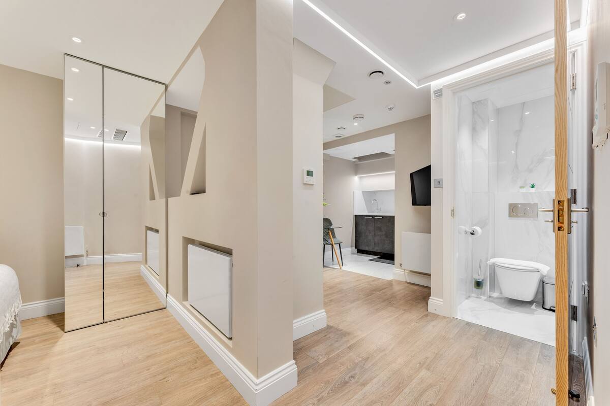 A modern hotel room with a large mirror, wooden flooring, a separate bathroom, and a kitchenette.