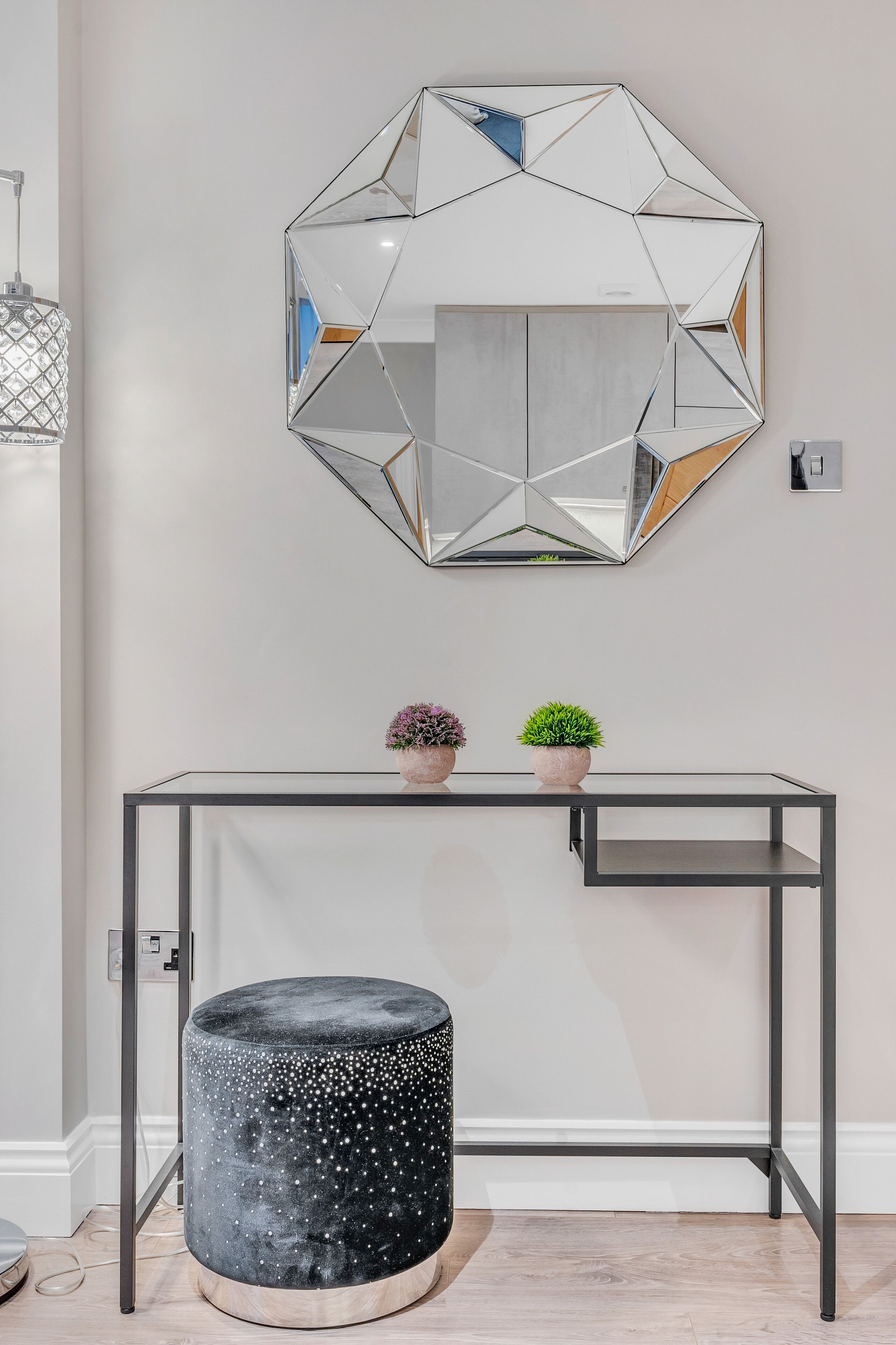 A modern room with a geometric mirror, a black console table, two potted plants, and a round ottoman.