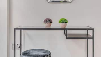 A modern room with a geometric mirror, a black console table, two potted plants, and a round ottoman.
