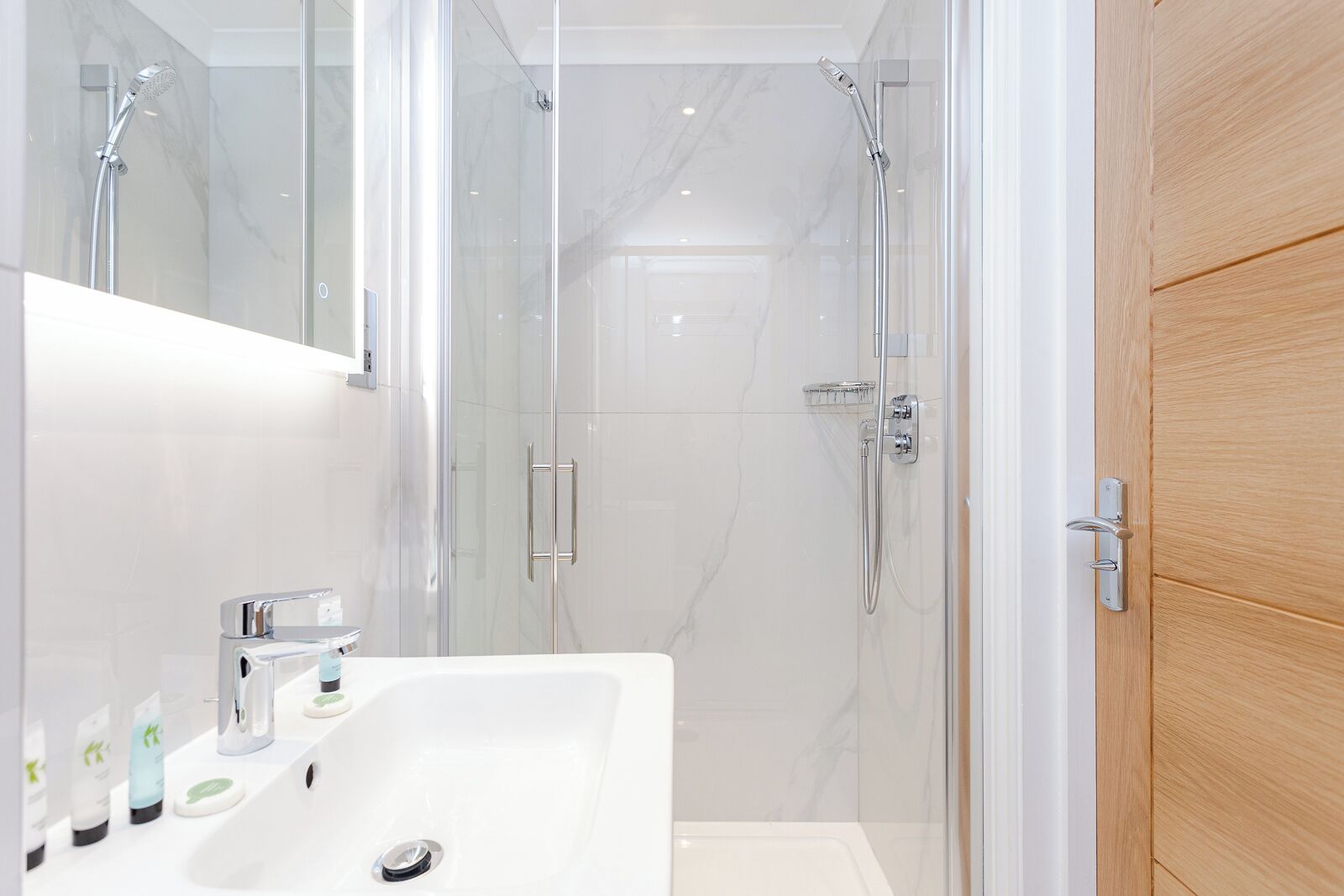 Une salle de bain moderne avec une douche à parois de verre, un lavabo blanc agrémenté d’articles de toilette et une porte en bois.