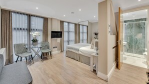 A modern hotel room with a bed, a TV, a glass table with chairs, a bathroom with a glass shower, and a large window with curtains. - Cleveland Residences Farringdon (London)