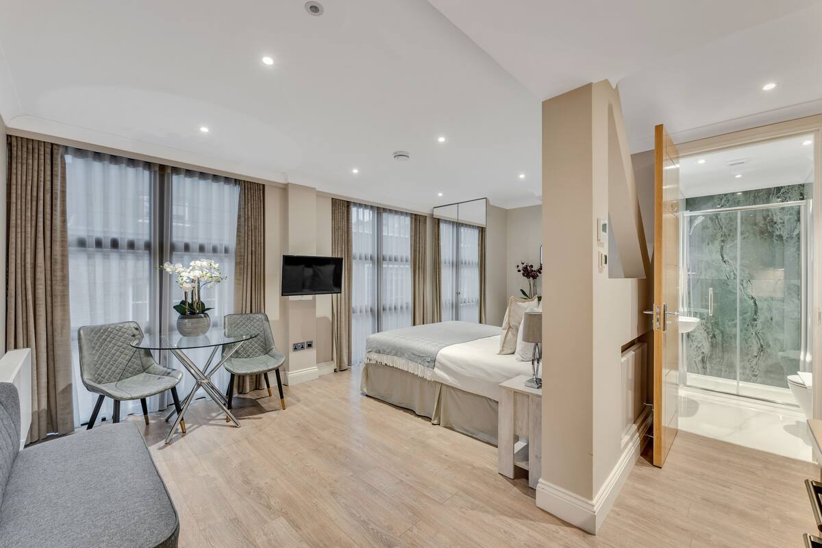 A modern hotel room with a bed, a TV, a glass table with chairs, a bathroom with a glass shower, and a large window with curtains.