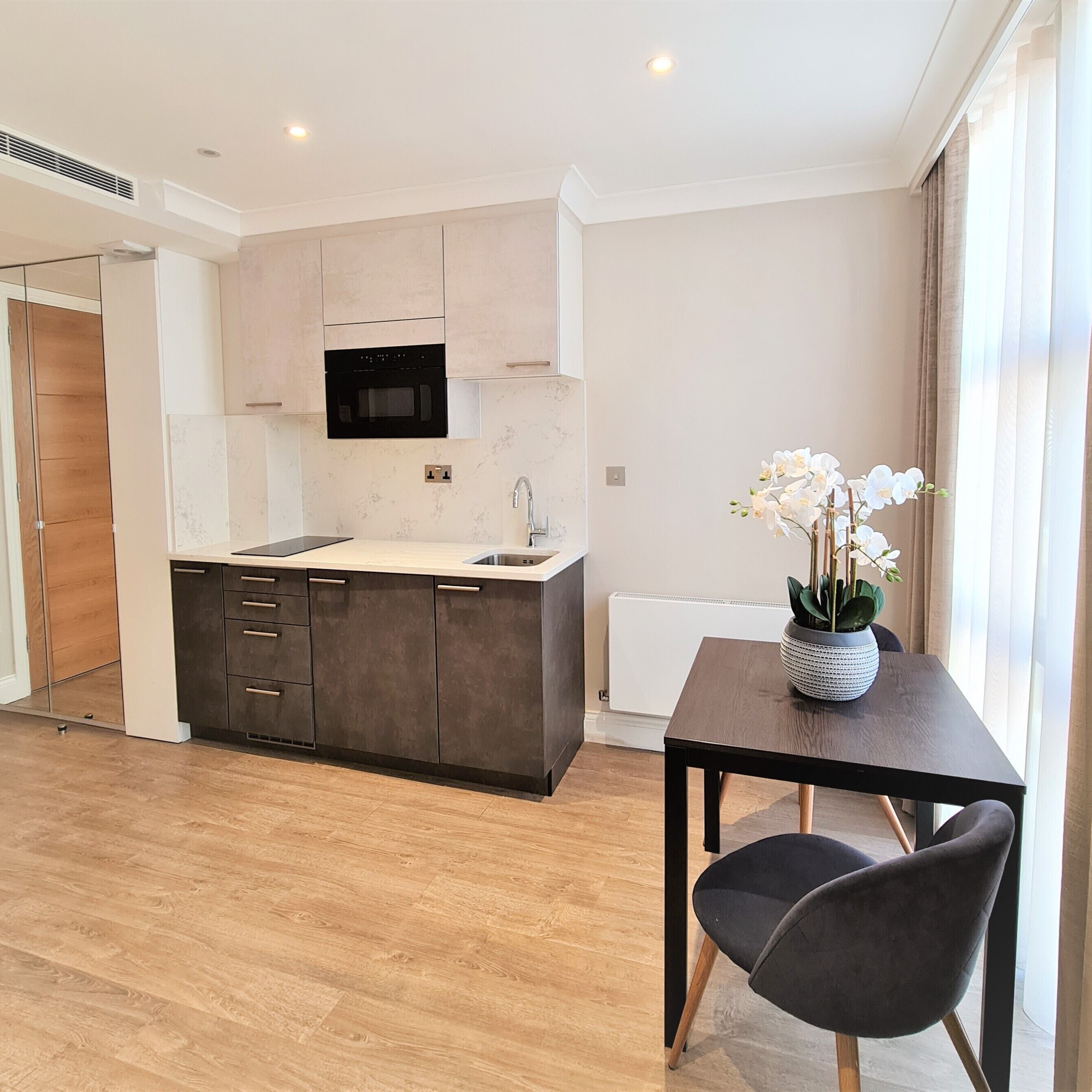 Una cocina moderna con armarios oscuros, microondas integrado y un fregadero. Hay una mesa de comedor con sillas y un jarrón de flores blancas.