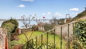 A garden with a metal railing, a stone wall, and a view of a body of water and a bridge in the distance.