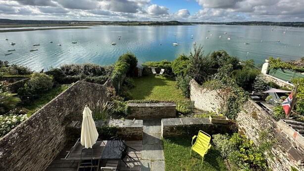 A terrace with stone walls, a table, chairs, and an umbrella overlooking a lake with boats.
