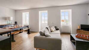 A modern living room with a sofa, a dining table, and a foosball table. There are windows providing natural light and a view of the outdoors.