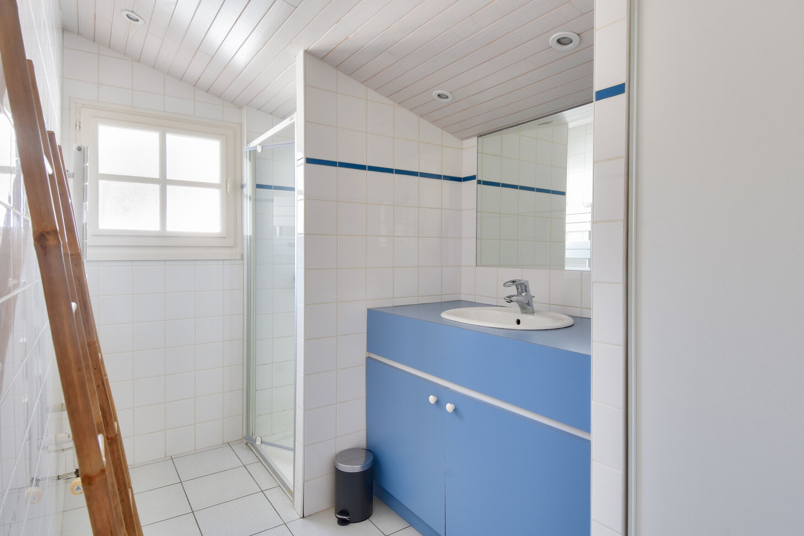 Een badkamer met een vloer van witte tegels, een blauwe wastafel met wastafel en spiegel, een douchegedeelte en een raam.
