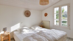Quarto com cama, mesa de cabeceira, luminária, um tapete de parede e janela com vista para o verde.