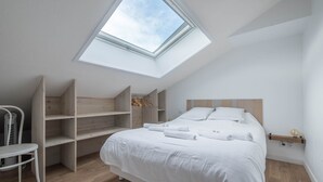 Un dormitorio moderno con tragaluz, cama con ropa de cama blanca, estanterías de madera y una mesita.