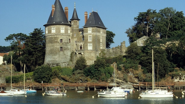 Un castillo histórico con múltiples torres, ubicado junto a un río tranquilo donde hay varias embarcaciones amarradas.