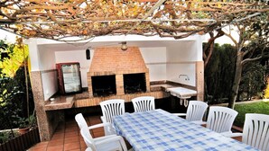 Une terrasse extérieure avec un barbecue en briques, des chaises blanches et une nappe à carreaux.