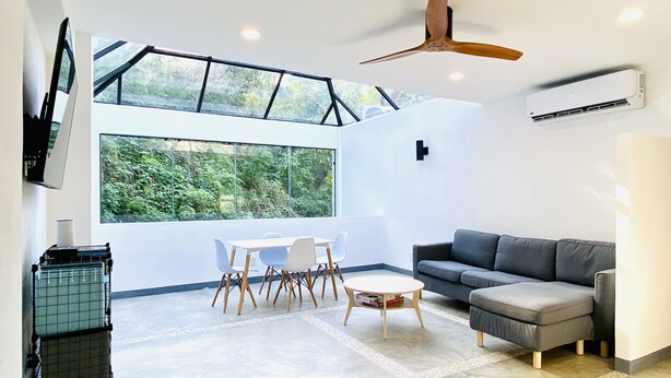 A modern living room with a large window, a ceiling fan, a grey sofa, and a dining area with white chairs and a table.