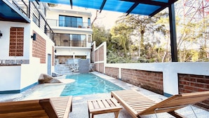 A rooftop pool area with a view of a modern building, wooden lounge chairs, and a covered patio.
