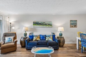 A living room with a dark blue sofa, two lamps, a coffee table with a beach design, and a painting of a coastal scene. - Heaven's Nest -- First Floor Duck Condo with direct access to Beach and Pool (Kitty Hawk)