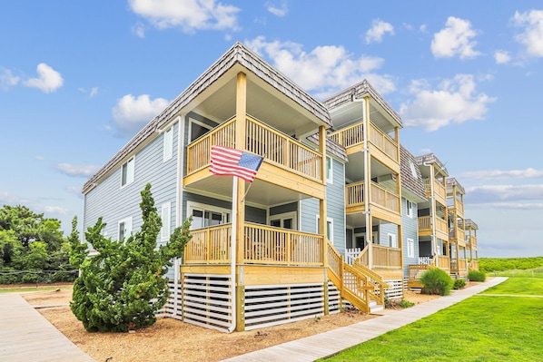 A multi-story residential building with balconies, a flag, and a well-maintained lawn. - Heaven's Nest -- First Floor Duck Condo with direct access to Beach and Pool (Kitty Hawk)