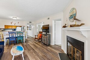 A living room with a fireplace, a dining area with chairs and a table, and a kitchen in the background. - Heaven's Nest -- First Floor Duck Condo with direct access to Beach and Pool (Kitty Hawk)