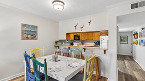 Un comedor con una mesa blanca, sillas coloridas y una cocina al fondo.