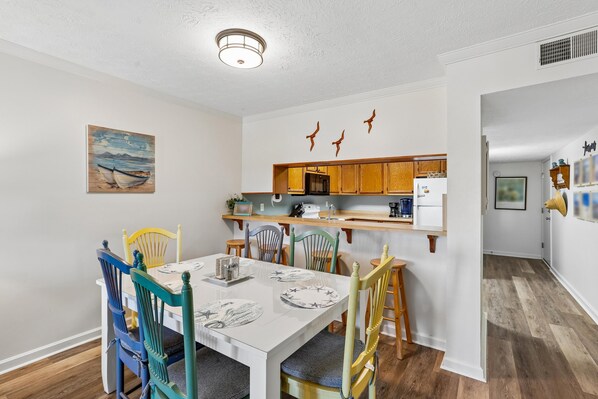 A dining area with a white table, colorful chairs, and a kitchen in the background. - Heaven's Nest -- First Floor Duck Condo with direct access to Beach and Pool (Kitty Hawk)