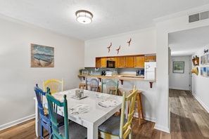 A dining area with a white table, colorful chairs, and a kitchen in the background. - Heaven's Nest -- First Floor Duck Condo with direct access to Beach and Pool (Kitty Hawk)